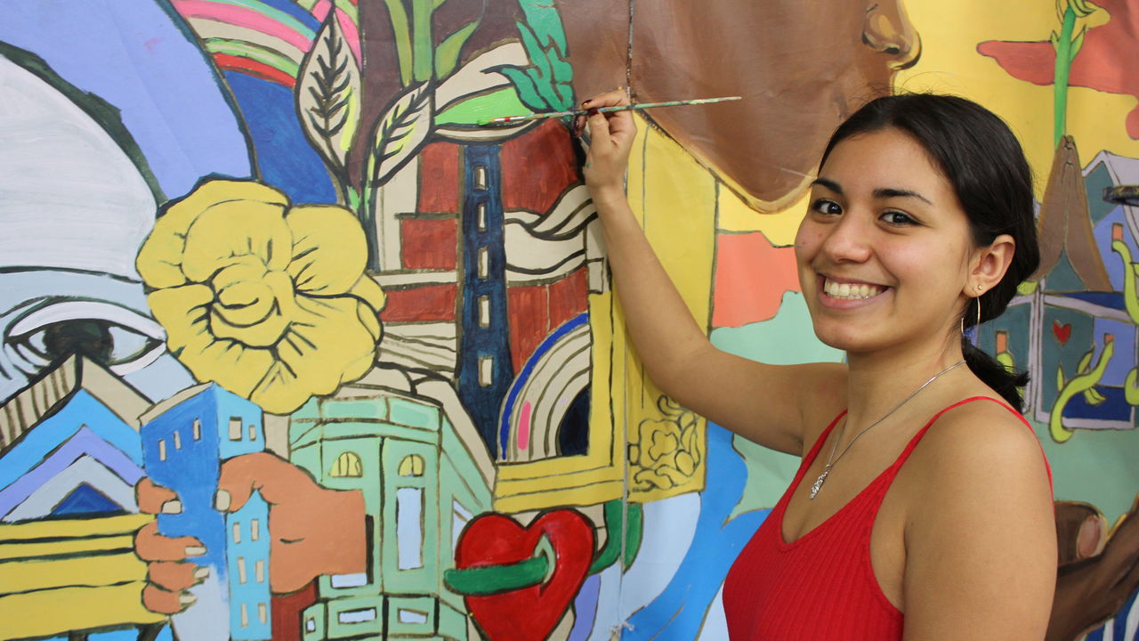 A youth artist holds a paintbrush to a colorful mural.
