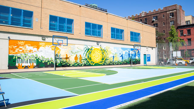Completed mural in school yard with bright colors, natural elements, figures playing and walking, a big sun in the middle and hands playing a keyboard. 
