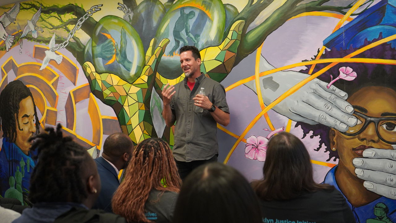People sitting and listening to a presenter speak in front of a mural. 