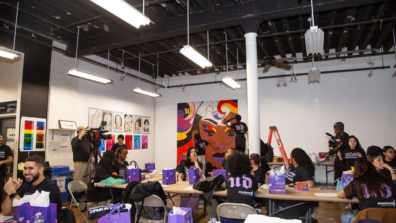 Wide photo of room, showcasing mural being painted, and tables set up with people talking and working. 