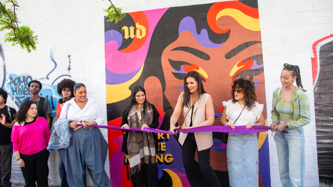 Group of people standing in front of a mural doing a ribbon cutting ceremony. 