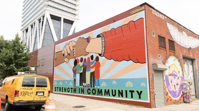 Mural that shows a group of people facing away from us with arms around each other, two bigger hands above the group holding each other's wrists, and the saying "strength in community" on the bottom. 