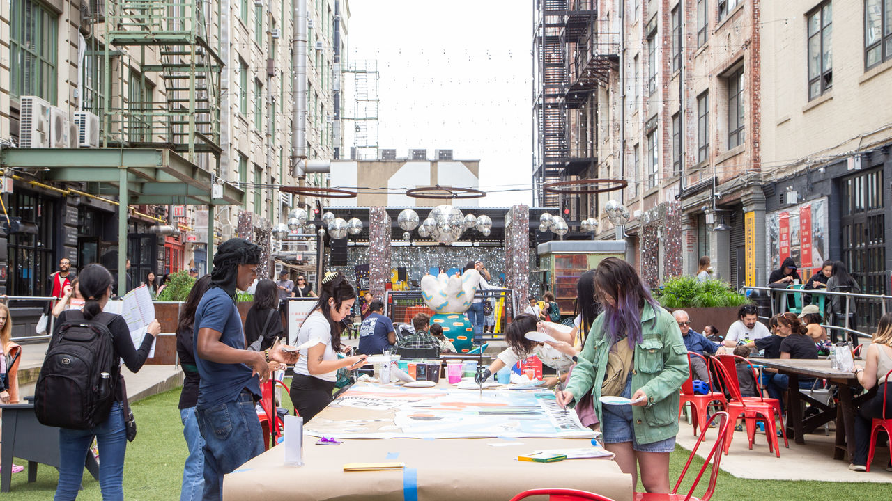 Wide photo showing people around a table painting a mural in a public courtyard. 