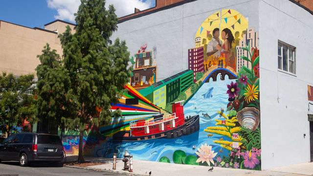 Mural showing a tugboat in water with rainbow rays, a couple dancing in a cityscape and natural elements. 