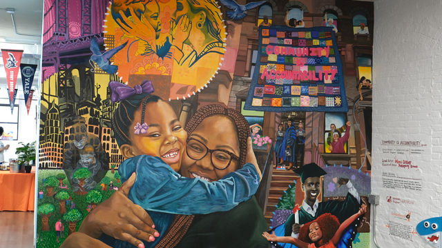 Mural featuring a mom and daughter hugging, another mom and child holding faces, a quilt that says "community is accountability, Brooklyn landmarks and a townhouse, a mom graduating and a child dressed as a butterfly