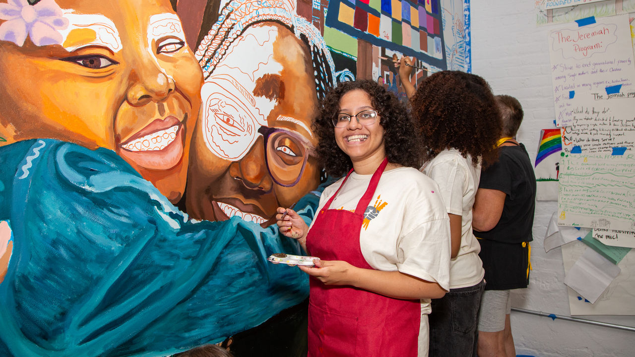A student holding a paint brush and pallet smiling next to her artwork 