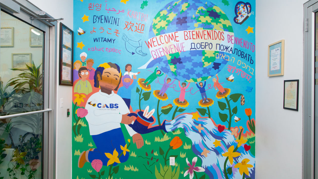 A mural showing a person in a CABS shirt watering plants with a group of people behind them, another group of people holding up a globe that has "welcome" written in multiple languages, and natural elements.